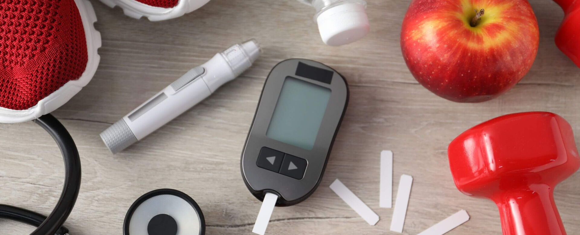 Fitnessgeräte und Diabetesmanagement: Trainingsschuhe, Apfel, Blutzuckermessgerät und Gewichte auf einem Tisch.